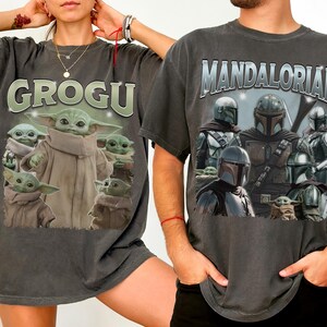 May include: Two dark gray t-shirts featuring Star Wars characters. One shirt displays "GROGU" above an image of Grogu. The other shirt has "MANDALORIAN" above images of Mandalorian characters and Grogu.