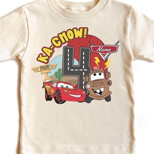 May include: Cream-colored t-shirt featuring a graphic of Lightning McQueen and Mater from the movie Cars. The design includes the text "Ka-Chow!" and a large number "4", with space for a name. A fun birthday shirt.
