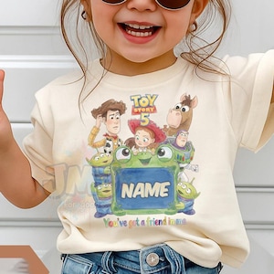 May include: A cream-coloured short-sleeve t-shirt featuring a colourful graphic print of Toy Story 5 characters including Woody, Jessie, Buzz Lightyear, and Bullseye surrounding a green frog holding a sign with the customisable text NAME.