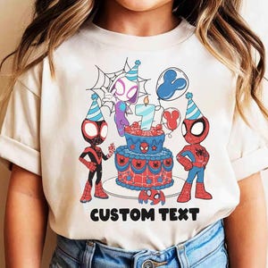 Op de afbeelding: Een beige t-shirt met een verjaardagstaartontwerp met het nummer 7, Spider-Man-personages en ballonnen. De woorden "CUSTOM TEXT" staan eronder in het zwart gedrukt. Het t-shirt wordt door een kind gedragen.