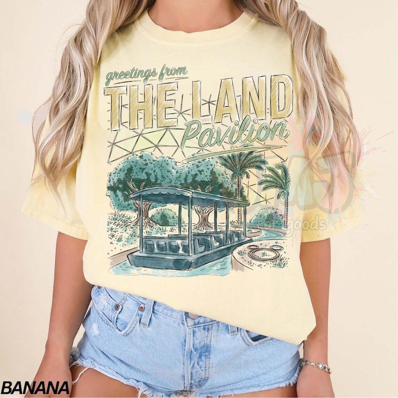 Puede incluir: Una camiseta de manga corta de color amarillo p&aacute;lido con un estampado gr&aacute;fico que incluye el texto Greetings from THE LAND Pavilion. El dise&ntilde;o muestra un barco en un canal, palmeras y una estructura de c&uacute;pula geod&eacute;sica en tonos verde azulado y beige.