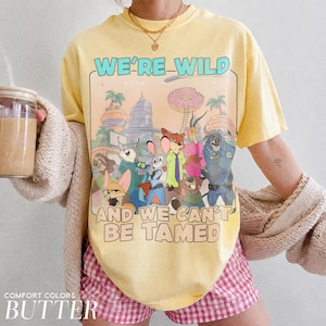K&ouml;nnte beinhalten: Ein buttergelbes Kurzarm-T-Shirt mit einem Grafikdruck verschiedener animierter Tierfiguren aus Zootopia. Der Text WE'RE WILD AND WE CAN'T BE TAMED ist in fetter Schrift oberhalb und unterhalb der farbenfrohen Zeichentrickillustration gedruckt.