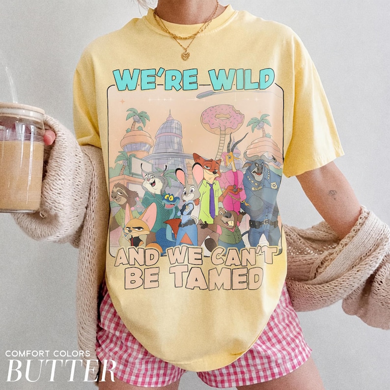 K&ouml;nnte beinhalten: Ein buttergelbes Kurzarm-T-Shirt mit einem Grafikdruck verschiedener animierter Tierfiguren aus Zootopia. Der Text WE'RE WILD AND WE CAN'T BE TAMED ist in fetter Schrift oberhalb und unterhalb der farbenfrohen Zeichentrickillustration gedruckt.
