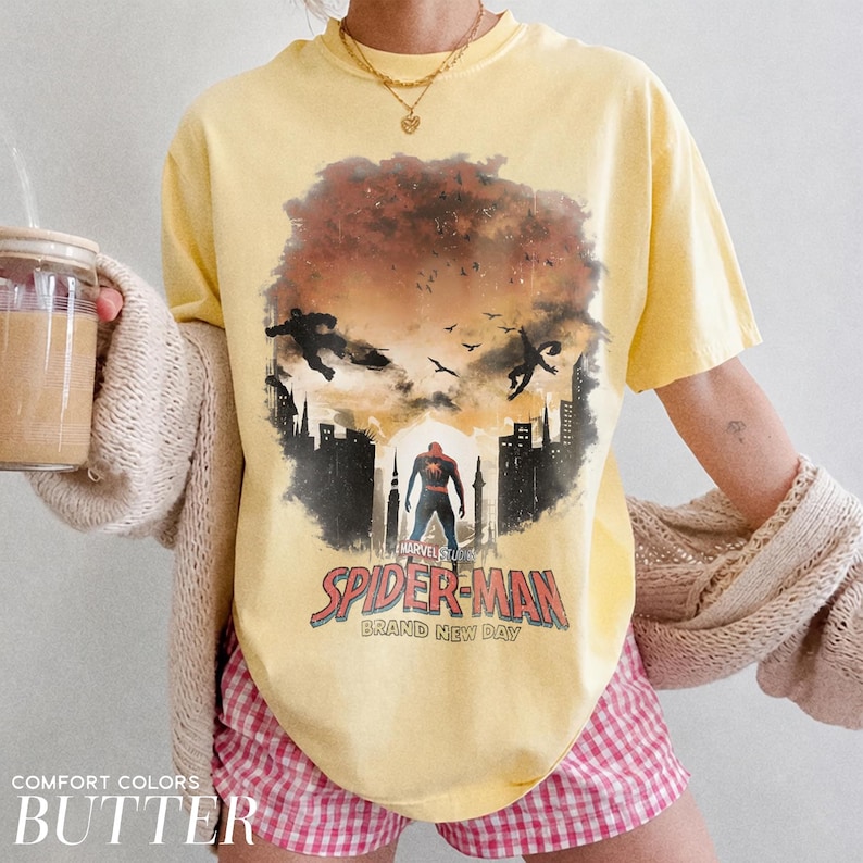 Pu&ograve; includere: Una t-shirt a maniche corte color giallo pallido con una stampa grafica di Spider-Man in piedi in una silhouette cittadina con due figure che saltano nel cielo. Il testo recita Spider-Man Brand New Day con un logo Marvel Studios.