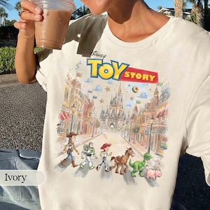 May include: Ivory-colored t-shirt featuring the Disney Toy Story logo and a colorful graphic of the characters walking down a street. The graphic includes Woody, Buzz Lightyear, Jessie, Bullseye, Rex, and Hamm. The shirt is a crew neck.