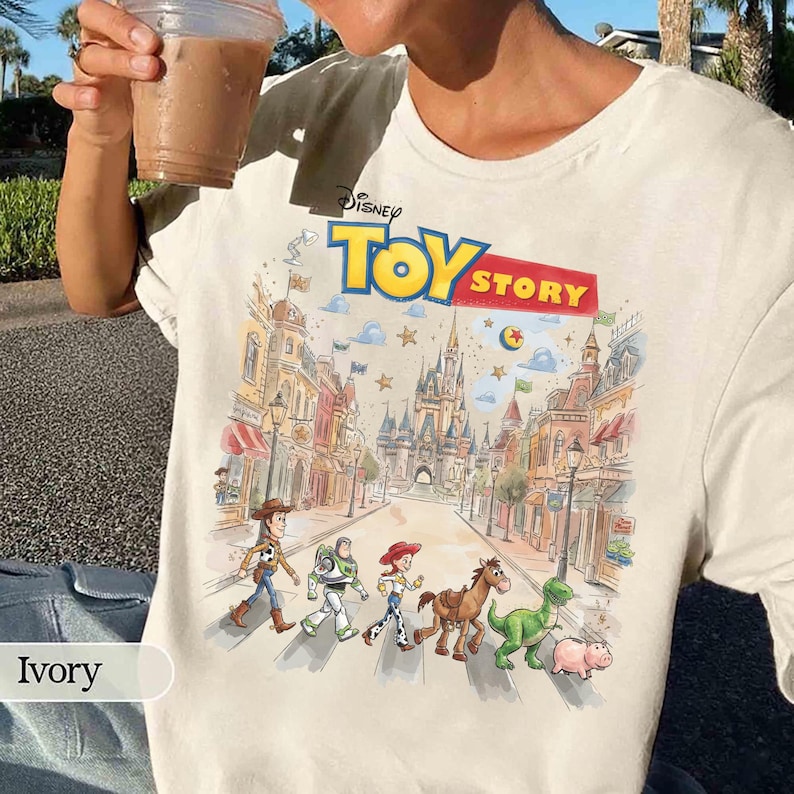 May include: Ivory-colored t-shirt featuring the Disney Toy Story logo and a colorful graphic of the characters walking down a street. The graphic includes Woody, Buzz Lightyear, Jessie, Bullseye, Rex, and Hamm. The shirt is a crew neck.