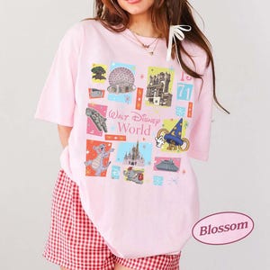 May include: A light pink t-shirt featuring a colorful graphic of Walt Disney World icons. The design includes images of the Spaceship Earth, Cinderella Castle, and other park attractions. The text "Walt Disney World" is also displayed.