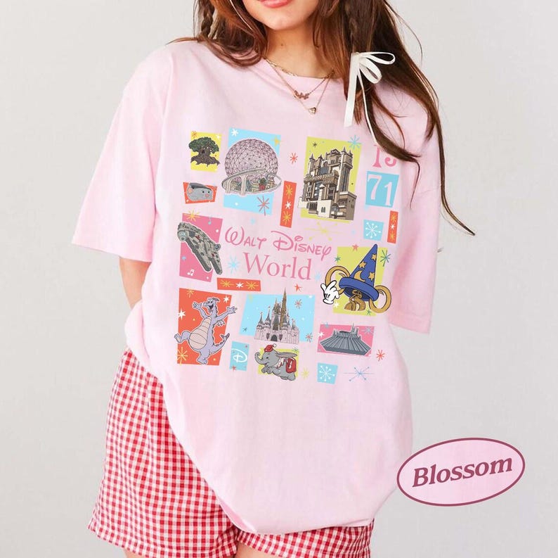 May include: A light pink t-shirt featuring a colorful graphic of Walt Disney World icons. The design includes images of the Spaceship Earth, Cinderella Castle, and other park attractions. The text "Walt Disney World" is also displayed.