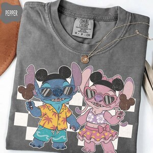 May include: A gray Comfort Colors t-shirt featuring a cartoon illustration of Stitch and Angel, each holding a Mickey Mouse-shaped ice cream. Stitch wears sunglasses and a Hawaiian shirt. Angel wears a skirt and sunglasses. A gold necklace is draped across the shirt.