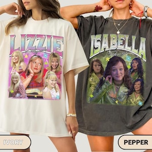 May include: Two oversized t-shirts featuring graphic prints. The ivory shirt displays the text Lizzie McGuire with multiple images of a blonde character. The pepper-colored shirt features the text Isabella Parigi with images of a brunette character.