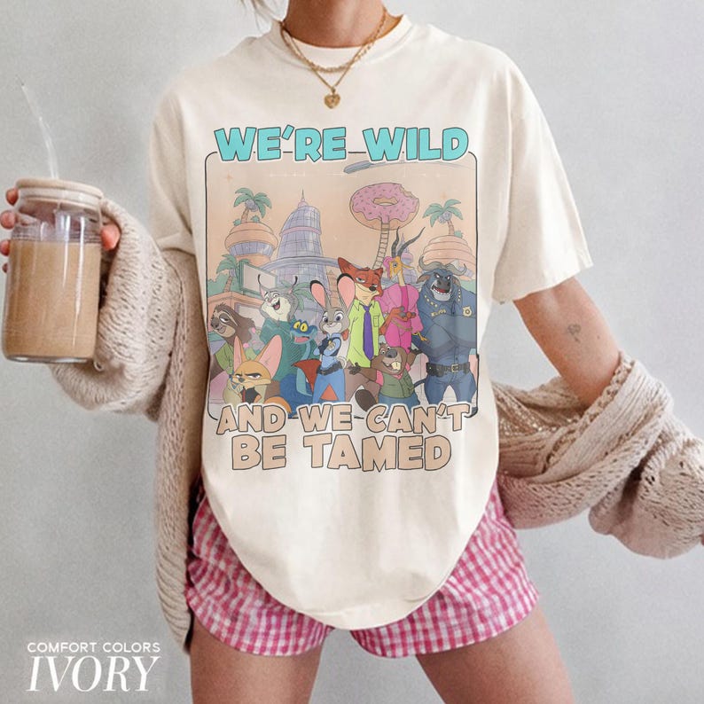 K&ouml;nnte beinhalten: Ein elfenbeinfarbenes Kurzarm-T-Shirt mit einem Grafikdruck verschiedener animierter Tierfiguren aus Zootopia. Der Text WE'RE WILD AND WE CAN'T BE TAMED ist in Blau und Beige gedruckt. Das T-Shirt wird mit einer beigen Strickjacke und rosa Vichy-Shorts getragen.