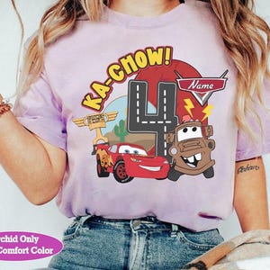 May include: A light purple t-shirt featuring a graphic of Lightning McQueen and Mater from the movie Cars, with the text "KA-CHOW!" and the number "4". The shirt also includes the text "Name" and "Orchid Only With Comfort Color".