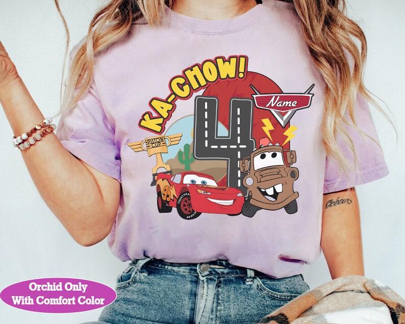 May include: A light purple t-shirt featuring a graphic of Lightning McQueen and Mater from the movie Cars, with the text "KA-CHOW!" and the number "4". The shirt also includes the text "Name" and "Orchid Only With Comfort Color".