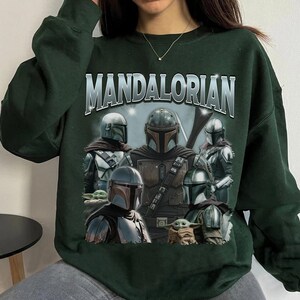 May include: Dark green sweatshirt featuring a graphic of characters from the Mandalorian series. The word "MANDALORIAN" is printed across the top in silver lettering. The graphic includes several figures in Mandalorian armor and a small green creature.