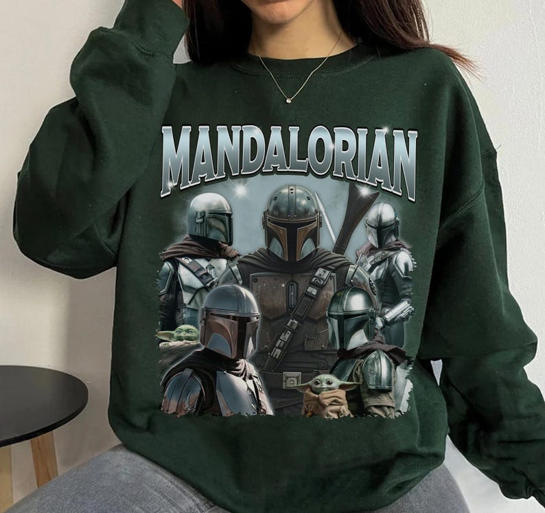 May include: Dark green sweatshirt featuring a graphic of characters from the Mandalorian series. The word "MANDALORIAN" is printed across the top in silver lettering. The graphic includes several figures in Mandalorian armor and a small green creature.