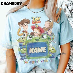 May include: A light blue t-shirt featuring a graphic print of Toy Story characters including Woody, Jessie, Bullseye, Buzz Lightyear, and three aliens. A large green frog holds a sign with the word NAME and the text You've got a friend in me.