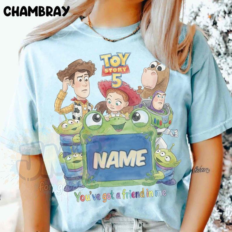 May include: A light blue t-shirt featuring a graphic print of Toy Story characters including Woody, Jessie, Bullseye, Buzz Lightyear, and three aliens. A large green frog holds a sign with the word NAME and the text You've got a friend in me.