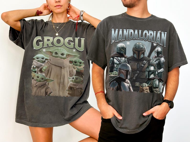 May include: Two dark gray t-shirts featuring Star Wars characters. One shirt displays "GROGU" above an image of Grogu. The other shirt has "MANDALORIAN" above images of Mandalorian characters and Grogu.