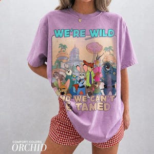 K&ouml;nnte beinhalten: Ein hellviolettes Kurzarm-T-Shirt mit einem Grafikdruck verschiedener animierter Tierfiguren aus Zootopia. Der Text WE'RE WILD AND WE CAN'T BE TAMED ist in Hellblau und Beige gedruckt. Das T-Shirt hat einen Rundhalsausschnitt und eine lockere Passform.