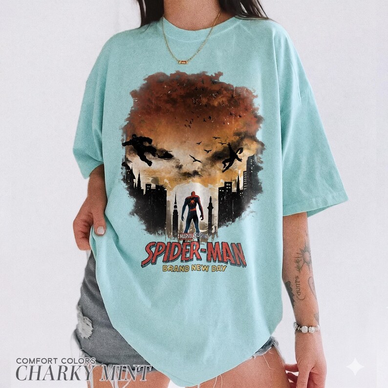 Pu&ograve; includere: Una t-shirt verde menta chiaro con una stampa grafica di Spider-Man in piedi in un paesaggio urbano con sagome di figure in un cielo arancione scuro. Il testo sotto la grafica recita Spider-Man Brand New Day in lettere rosse e nere.