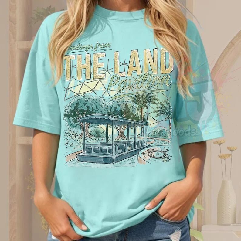 Puede incluir: Una camiseta de manga corta de color azul claro con un estampado gr&aacute;fico que incluye el texto Greetings from THE LAND Pavilion. El dise&ntilde;o muestra un barco estilizado, palmeras y una estructura de c&uacute;pula geom&eacute;trica en tonos azul, verde y amarillo.