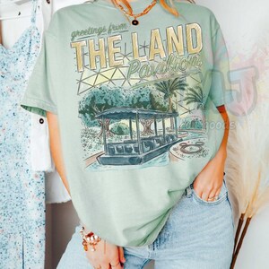 Puede incluir: Una camiseta de manga corta de color verde claro con un estampado gr&aacute;fico que incluye el texto 'greetings from THE LAND Pavilion'. La ilustraci&oacute;n muestra un barco sobre el agua, palmeras y una estructura de c&uacute;pula geod&eacute;sica.