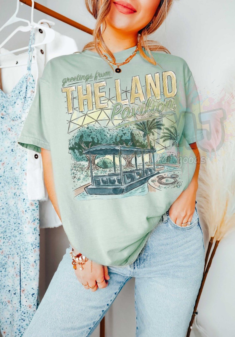 Puede incluir: Una camiseta de manga corta de color verde claro con un estampado gr&aacute;fico que incluye el texto 'greetings from THE LAND Pavilion'. La ilustraci&oacute;n muestra un barco sobre el agua, palmeras y una estructura de c&uacute;pula geod&eacute;sica.