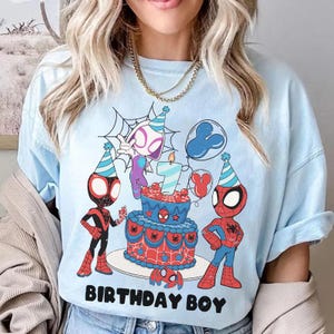 Op de afbeelding: Lichtblauw T-shirt met een kleurrijke afbeelding van Spider-Man-personages, een verjaardagstaart en ballonnen. De taart heeft een kaars met het cijfer 7. De tekst "BIRTHDAY BOY" staat onderaan. Een casual kledingstuk.