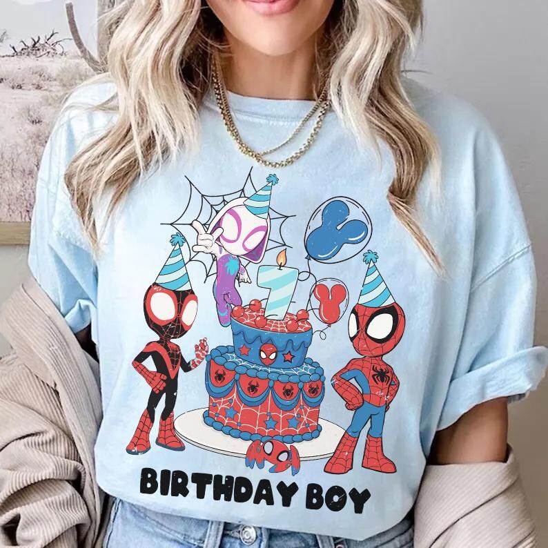 Op de afbeelding: Lichtblauw T-shirt met een kleurrijke afbeelding van Spider-Man-personages, een verjaardagstaart en ballonnen. De taart heeft een kaars met het cijfer 7. De tekst "BIRTHDAY BOY" staat onderaan. Een casual kledingstuk.