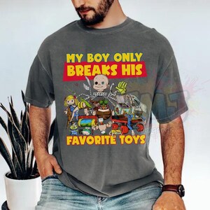 May include: A charcoal gray short-sleeve t-shirt featuring a graphic print of various toy characters and the yellow text "MY BOY ONLY BREAKS HIS FAVORITE TOYS" in a red box. The design includes a doll head, a green hand, and other toy figures.