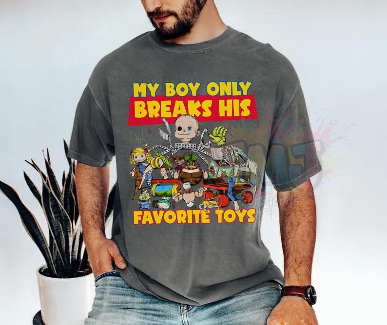 May include: A charcoal gray short-sleeve t-shirt featuring a graphic print of various toy characters and the yellow text "MY BOY ONLY BREAKS HIS FAVORITE TOYS" in a red box. The design includes a doll head, a green hand, and other toy figures.