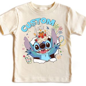 K&ouml;nnte beinhalten: Cremefarbenes T-Shirt mit einem blauen Cartoon-Charakter, der einen Nachtisch auf dem Kopf tr&auml;gt und aus einem zerrissenen Papiereffekt auftaucht. Der Text "CUSTOM Text" befindet sich &uuml;ber der Figur. Bunte Feuerwerke und Konfetti.