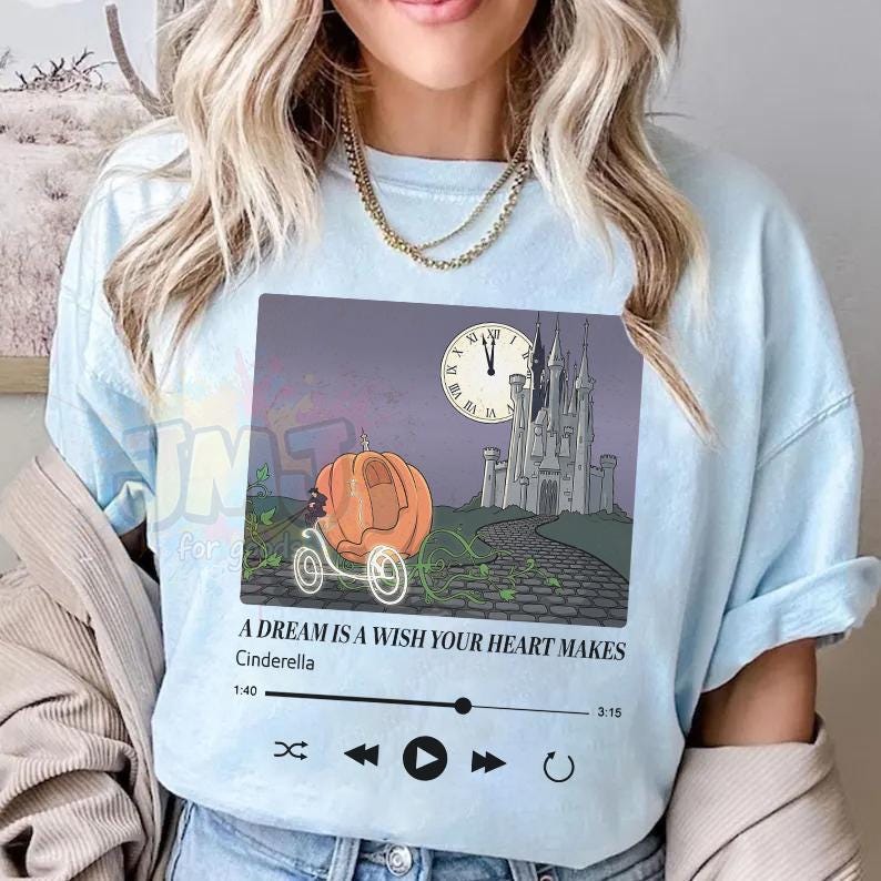May include: A light blue t-shirt featuring a graphic of a pumpkin carriage, a castle, and a clock face. Below the image, the text reads A DREAM IS A WISH YOUR HEART MAKES, Cinderella, 1:40, 3:15, and a digital music player interface with playback icons.