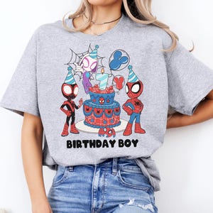 Op de afbeelding: Lichtgrijs t-shirt met een kleurrijke afbeelding van Spider-Man-personages, een verjaardagstaart en de tekst "BIRTHDAY BOY". De taart heeft een kaars met het nummer 7. Het ontwerp is rood, blauw en zwart.