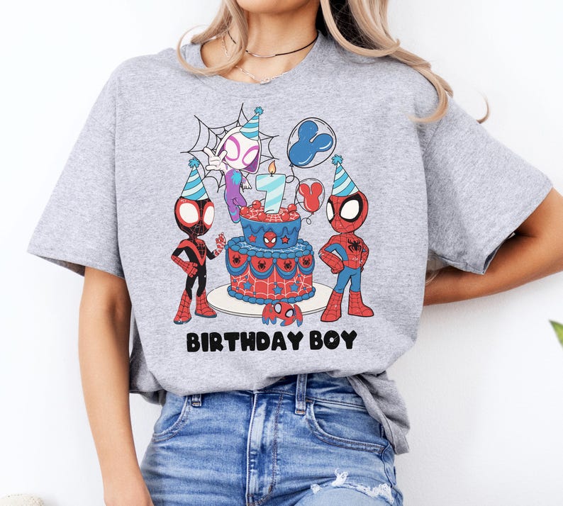 Op de afbeelding: Lichtgrijs t-shirt met een kleurrijke afbeelding van Spider-Man-personages, een verjaardagstaart en de tekst "BIRTHDAY BOY". De taart heeft een kaars met het nummer 7. Het ontwerp is rood, blauw en zwart.