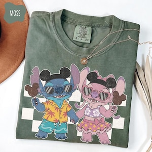 May include: Moss green t-shirt featuring a cartoon couple, each holding a Mickey Mouse ice cream bar. The characters wear sunglasses and summer attire. The shirt has a checkerboard pattern and a Comfort Colors tag.