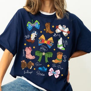 May include: Navy blue t-shirt featuring colorful cartoon bow designs and character names. The shirt includes images of cowboy boots, bows, and a space ranger. The text includes "Jessie", "Woody", "Buzz Lightyear", and "Bo Peep".