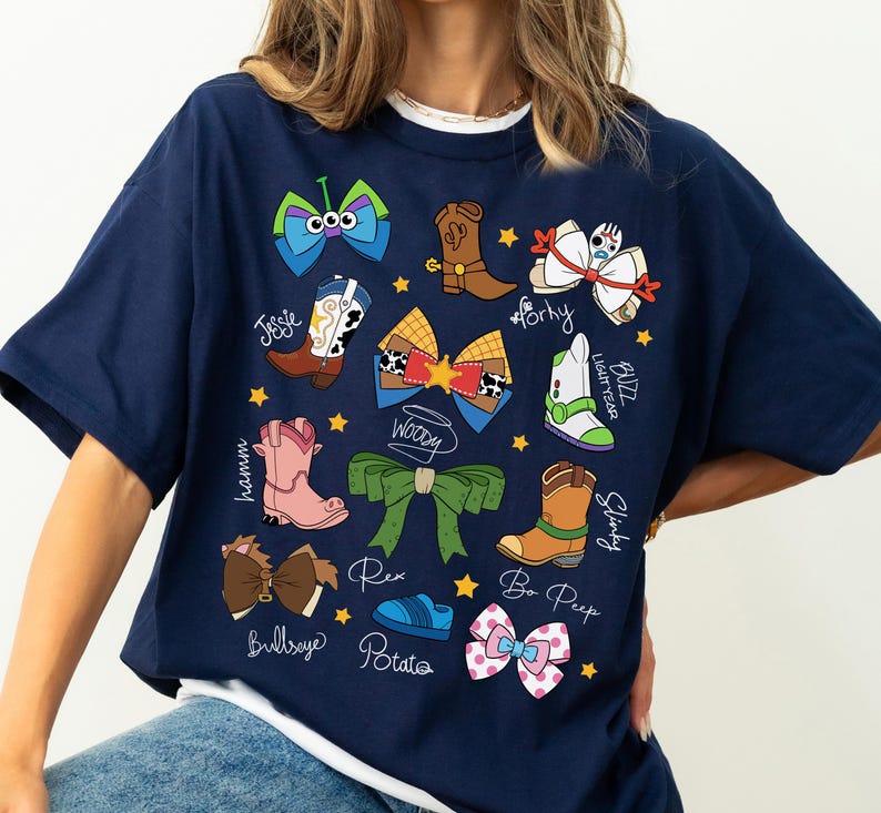 May include: Navy blue t-shirt featuring colorful cartoon bow designs and character names. The shirt includes images of cowboy boots, bows, and a space ranger. The text includes "Jessie", "Woody", "Buzz Lightyear", and "Bo Peep".