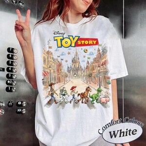 May include: White t-shirt featuring a colorful graphic of Toy Story characters crossing a street with a castle in the background. The Disney Toy Story logo is at the top. The shirt has the words "Comfort Colors White" on the bottom right.