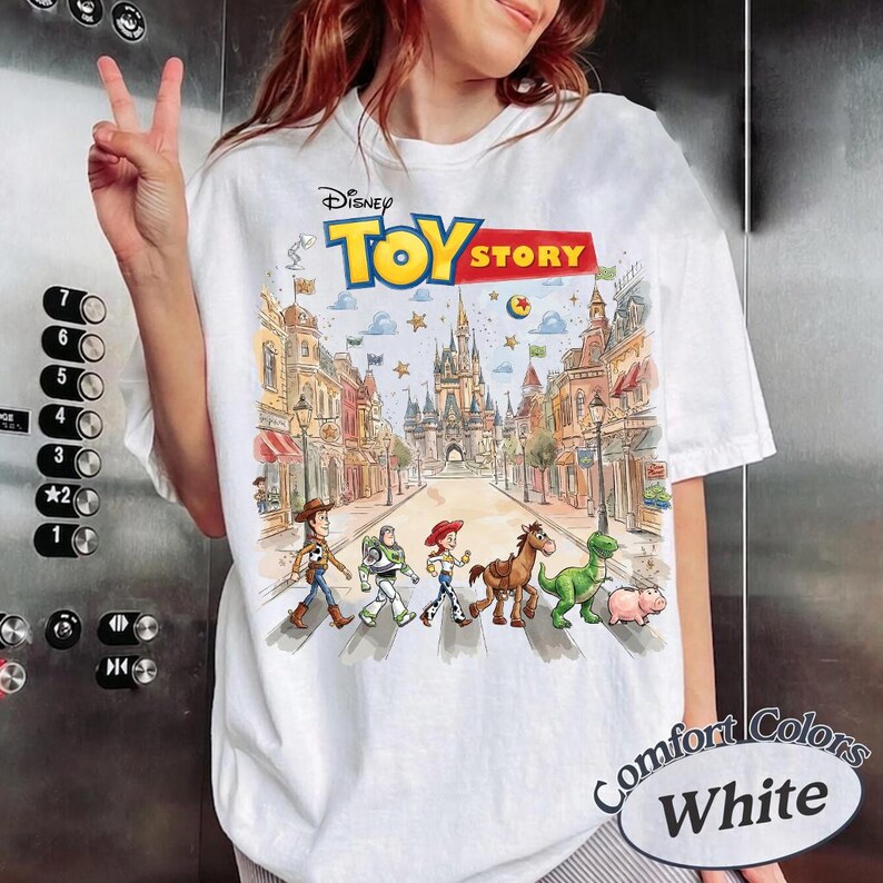 May include: White t-shirt featuring a colorful graphic of Toy Story characters crossing a street with a castle in the background. The Disney Toy Story logo is at the top. The shirt has the words "Comfort Colors White" on the bottom right.