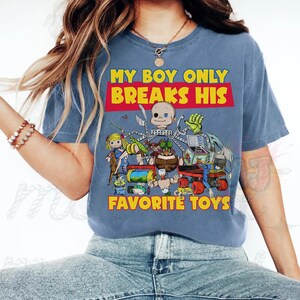 May include: A blue short-sleeved t-shirt featuring a graphic print of various toy characters and the text "MY BOY ONLY BREAKS HIS FAVORITE TOYS" in yellow and red. The design includes a doll, a green insect-like figure, and other mechanical toys.
