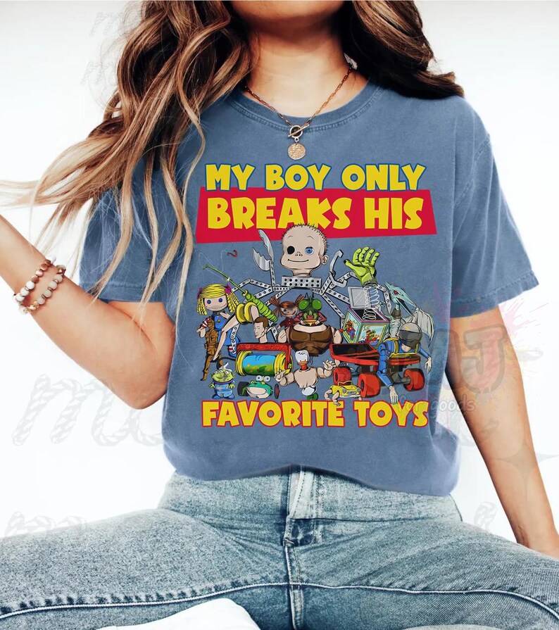 May include: A blue short-sleeved t-shirt featuring a graphic print of various toy characters and the text "MY BOY ONLY BREAKS HIS FAVORITE TOYS" in yellow and red. The design includes a doll, a green insect-like figure, and other mechanical toys.
