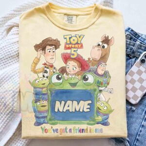 May include: A pale yellow t-shirt featuring a Toy Story 5 graphic with Woody, Jessie, Bullseye, Buzz Lightyear, and three green aliens surrounding a central green frog-shaped frame with the word NAME and the text You've got a friend in me.
