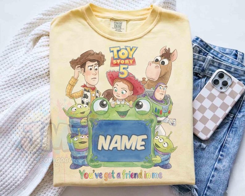 May include: A pale yellow t-shirt featuring a Toy Story 5 graphic with Woody, Jessie, Bullseye, Buzz Lightyear, and three green aliens surrounding a central green frog-shaped frame with the word NAME and the text You've got a friend in me.