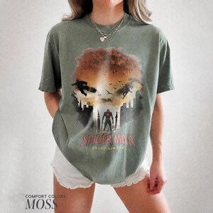 Pu&ograve; includere: Una t-shirt a maniche corte color verde muschio con una stampa grafica di Spider-Man in piedi in un paesaggio urbano con una grande esplosione arancione sullo sfondo. Il testo recita Spider-Man Brand New Day. La maglietta ha un girocollo.