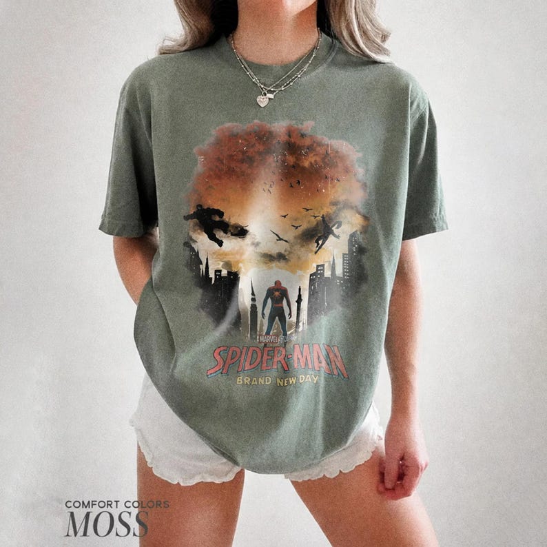 Pu&ograve; includere: Una t-shirt a maniche corte color verde muschio con una stampa grafica di Spider-Man in piedi in un paesaggio urbano con una grande esplosione arancione sullo sfondo. Il testo recita Spider-Man Brand New Day. La maglietta ha un girocollo.