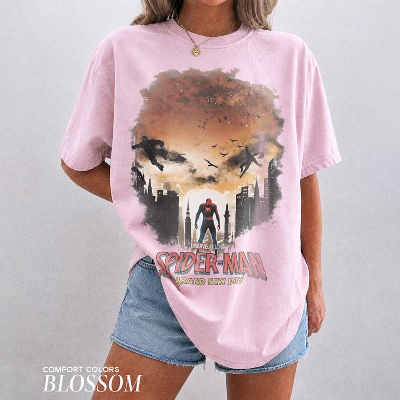 Pu&ograve; includere: Una t-shirt a maniche corte rosa chiaro con una stampa grafica di Spider-Man in un paesaggio urbano con sagome nel cielo. Il testo recita Marvel Studios Spider-Man Brand New Day in un carattere rosso e bianco dall'aspetto vissuto.