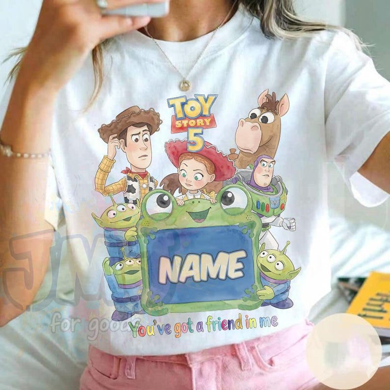 May include: A white t-shirt featuring a colourful graphic of Toy Story characters including Woody, Jessie, Buzz Lightyear, Bullseye, and green aliens surrounding a large green frog holding a blue sign with the text NAME and Toy Story 5.