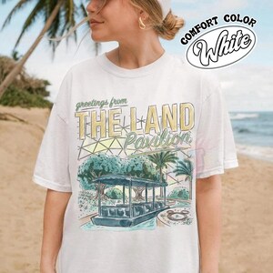 Puede incluir: Una camiseta blanca de manga corta con un estampado gr&aacute;fico que incluye el texto 'greetings from THE LAND Pavilion'. El dise&ntilde;o muestra un barco estilizado sobre el agua, palmeras y una estructura de c&uacute;pula geom&eacute;trica en verde, amarillo y azul.