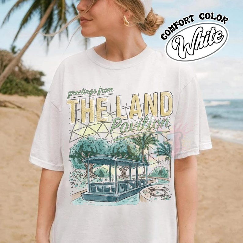 Puede incluir: Una camiseta blanca de manga corta con un estampado gr&aacute;fico que incluye el texto 'greetings from THE LAND Pavilion'. El dise&ntilde;o muestra un barco estilizado sobre el agua, palmeras y una estructura de c&uacute;pula geom&eacute;trica en verde, amarillo y azul.